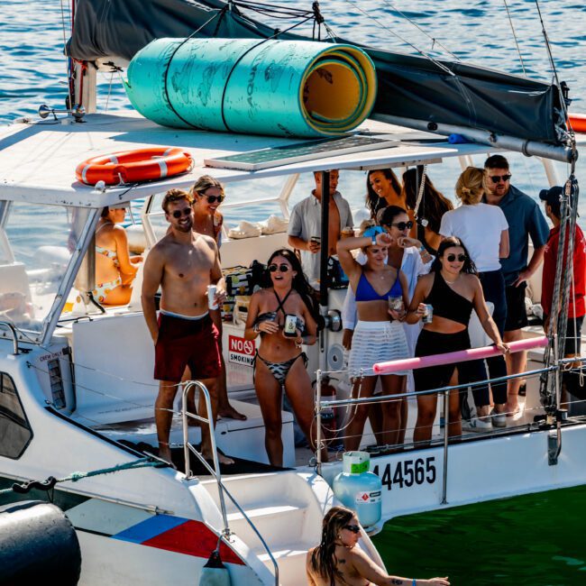 A group of people enjoying a sunny day on a boat, some holding drinks and others in swimwear. A few individuals are on the deck, while some are in the water near the boat. The Yacht Social Club Sydney Boat Hire ensures an unforgettable experience aboard their luxurious vessels.