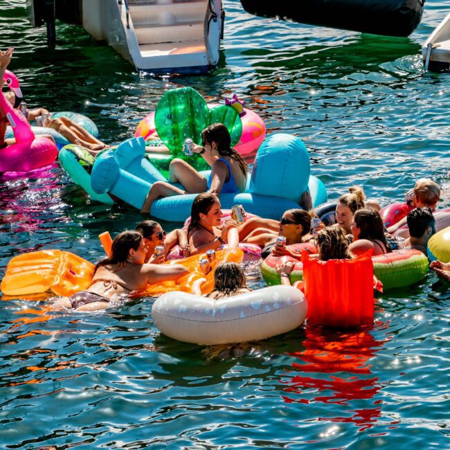 A group of people floating on colorful inflatable rafts enjoys a sunny day in the water near a boat. The clear water and cheerful atmosphere make for perfect socializing. The boat's stairway and name, "Italy St. Sydney," are visible in the background, showcasing the Luxury Yacht Rentals Sydney experience.