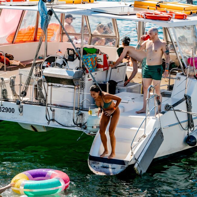 People enjoying a sunny day on a luxury yacht. A woman in a bikini prepares to jump into the clear water, where inflatable pool tubes float. Other people relax on the yacht, including a man standing on the side. The atmosphere is festive, perfect for Boat Rental and Parties Sydney The Yacht Social Club offers.