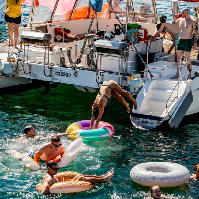 A group of friends enjoys a sunny day on the water, with several people swimming and floating on colorful inflatables near a sailboat. One person leaps into the water through a donut-shaped float, while others relax on the boat. The scene is lively and fun, perfect for The Yacht Social Club Event Boat Charters.