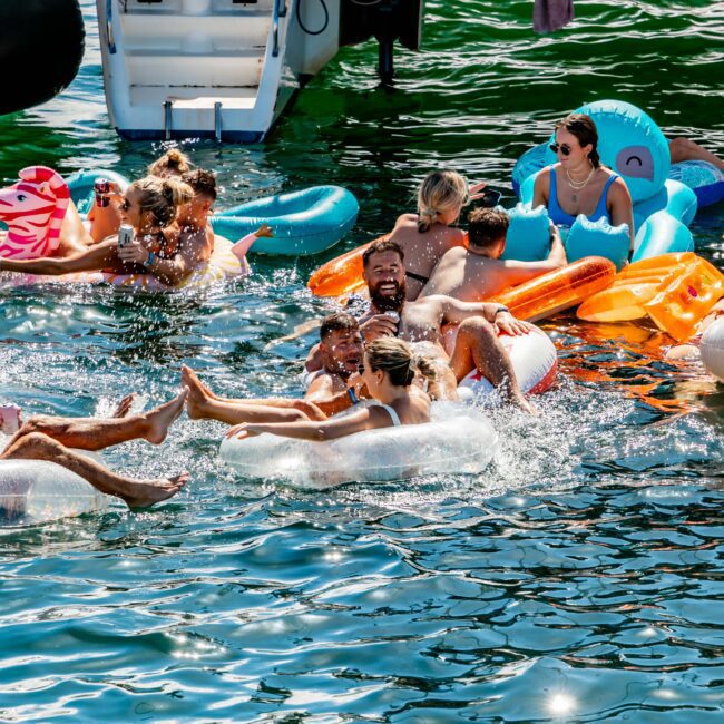 A group of people enjoying a sunny day on the water, floating on various inflatable rafts and tubes near a luxury yacht. Some are splashing in the water, while others relax on their inflatables. The atmosphere at this Yacht Social Club event is lively and joyful.