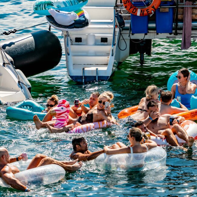 A group of people enjoying a sunny day on the water, floating on colorful inflatable tubes and rafts near a boat. Some are sitting or standing on the boat, while others relax in the clear, blue-green water. The boat's life ring reads "LADY S SYDNEY," indicating it's part of The Yacht Social Club Event.