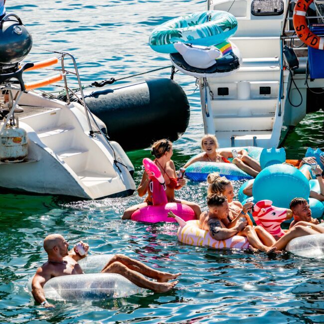 People enjoy a sunny day on the water, floating on various inflatable rafts including a flamingo and unicorn. Several boats from Sydney Harbour Boat Hire The Yacht Social Club are anchored nearby, with some participants lounging on the decks. The water is clear and blue, creating a vibrant, summery scene.