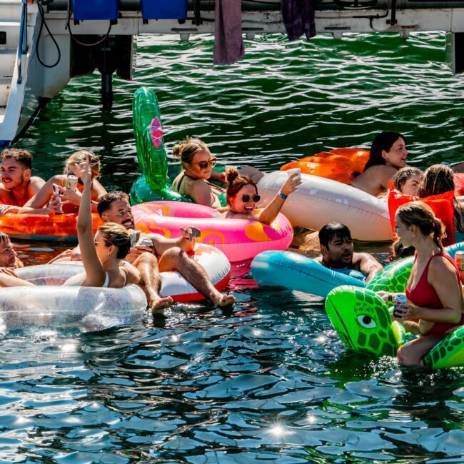 A group of people enjoys a sunny day on the water, lounging on colorful inflatable floats in shapes like a donut and a turtle. Smiling and chatting, they raise drinks close to a boat docked nearby with "Sydney" visible on life rings. It's all part of The Yacht Social Club event hosted by The Yacht Social Club Sydney Boat Hire.