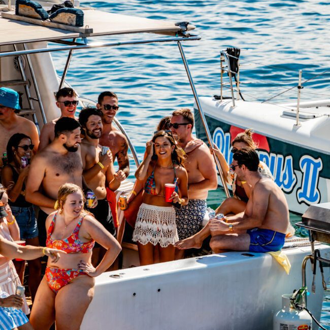 A group of people enjoying a sunny day on a boat named Olympus II, part of The Yacht Social Club Sydney Boat Hire. They are dressed in swimwear, holding drinks, and smiling. Anchored in blue waters, a woman in the center wears sunglasses and a floral skirt.