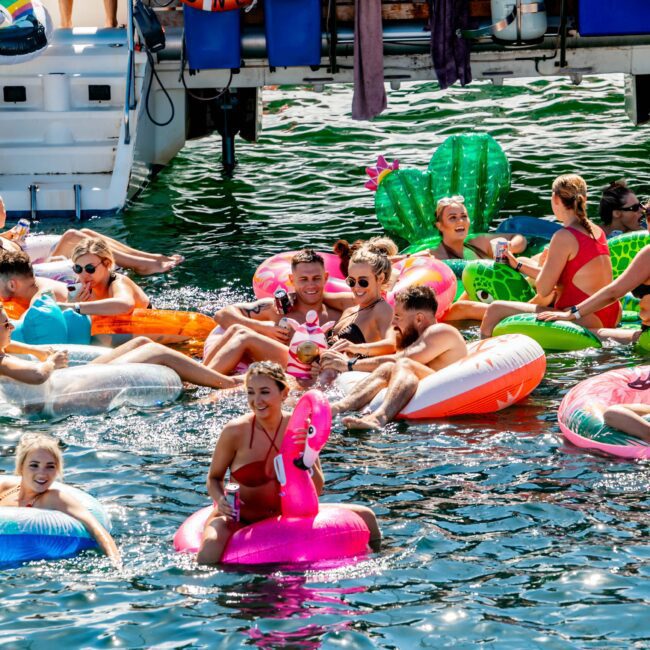 A lively group of people relax and enjoy the water on colorful inflatable rafts in a sunny outdoor setting. The water is a vibrant blue, and a boat with "Sydney City" written on life rings is docked nearby, hinting at services from Luxury Yacht Rentals Sydney. The atmosphere is festive and joyful.