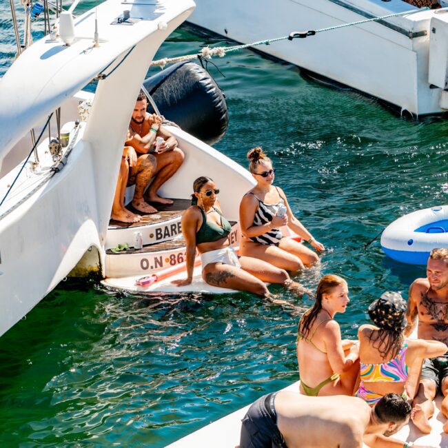 A group of people on a boat are enjoying a sunny day on the water. Some are sitting on the boat's steps, while others are in the water or on a floating mat, relaxing and having fun. This scene captures the essence of The Yacht Social Club Event Boat Charters and Sydney Harbour Boat Hire.