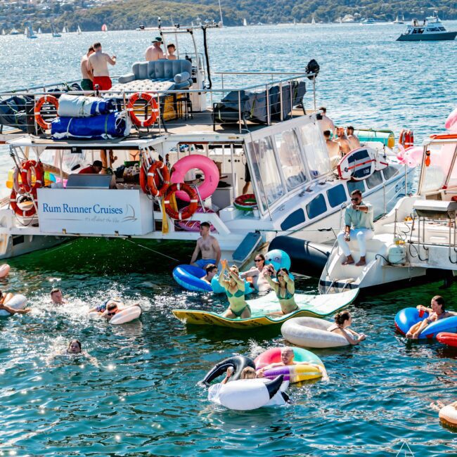A sunny scene of people enjoying a boat party. The boat, labeled "Rum Runner Cruises," is anchored while people in swimsuits play and float on inflatables in the water. Trees and hills are visible in the background, along with other boats from The Yacht Social Club Event Boat Charters in the distance.