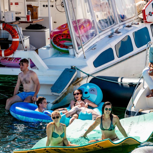 A group of people enjoy a sunny day on a lake, lounging on colorful inflatable floats and mats near a white boat. Some are sitting or lying down, soaking up the sun, while others are standing or swimming in the water. Everyone seems relaxed and happy at The Yacht Social Club Event Boat Charters.