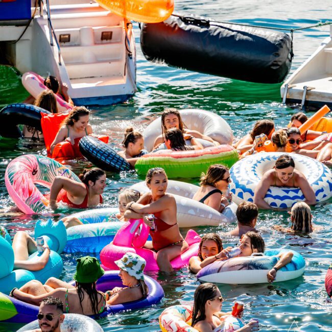 People on various inflatable rings and rafts are enjoying themselves in the water near a boat. Some are relaxing, while others are conversing and taking photos. Organized by The Yacht Social Club Sydney Boat Hire, the atmosphere appears lively and festive, suggesting a fun, social gathering.
