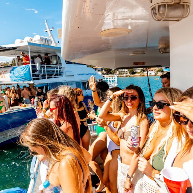 A group of people on a boat are enjoying a sunny day. Many are wearing swimwear and holding drinks. Some are taking photos, while others are dancing or chatting. Another boat from The Yacht Social Club Sydney Boat Hire can be seen in the background. The atmosphere is lively and festive.