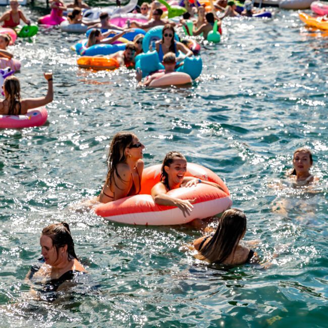 People enjoying a sunny day in the water, with some floating on inflatable pool toys and others swimming. Several boats from The Yacht Social Club Sydney Boat Hire are in the background, and the atmosphere appears lively and festive.