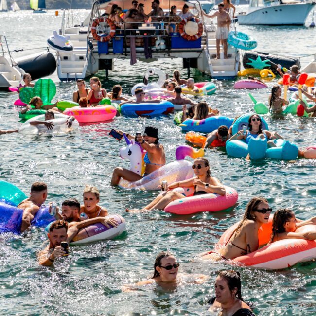 A lively scene of people on colorful inflatables enjoying a boat party in a sunny harbor. A luxury yacht rental from The Yacht Social Club in the background is filled with people, while others float and swim around, creating a festive atmosphere with a variety of inflatable toys and water activities.