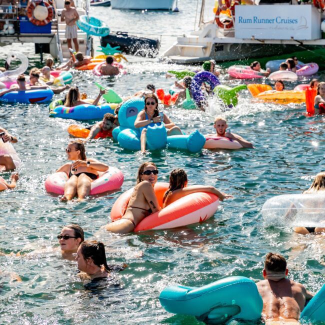 A lively scene of people swimming and relaxing on colorful inflatable floats in the ocean near anchored boats. The group includes various individuals enjoying the water, with some boats in the background, including one named "Rum Runner Cruises," during a Boat Parties Sydney The Yacht Social Club event.