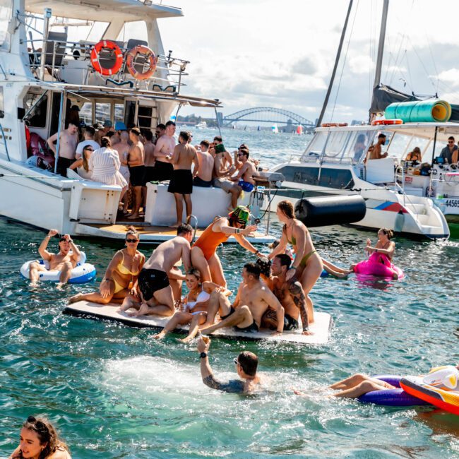 People are enjoying a sunny day on the water, some aboard a boat from The Yacht Social Club while others float nearby on inflatable rafts and a platform. The scene is lively, with socializing and playful activity. A bridge is visible in the background under a partly cloudy sky.