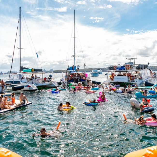 A crowded boat party with numerous people enjoying the water. Some are on inflatable floaties, while others swim or relax on boats nearby. The scene is lively with clear skies and several boats anchored in the background, hosted by The Yacht Social Club Sydney Boat Hire.