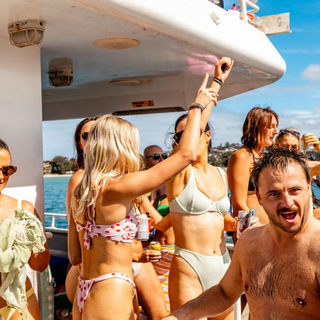 A group of people in swimwear are enjoying a lively party on a boat. Some are dancing, and others are holding drinks. The sun is shining against the clear sky and distant shoreline. Everyone appears to be having an amazing time with Sydney Harbour Boat Hire The Yacht Social Club.