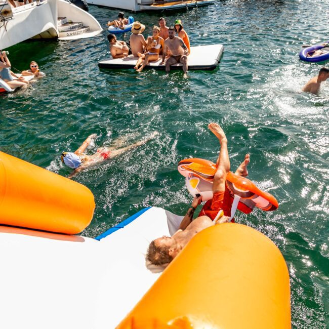 People are enjoying a sunny day on a lake with boats around. One person is mid-air on an inflatable slide, while others swim and relax on a floating mat. Some are floating on swim rings. The atmosphere resembles the fun of The Yacht Social Club Sydney Boat Hire events, with water reflecting the bright sunlight.
