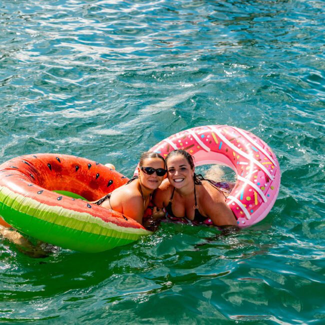Two women are smiling and enjoying the water while floating on colorful inflatable rings—one resembling a watermelon and the other a donut. Around them, other people on inflatables create a lively and fun atmosphere, reminiscent of boat parties hosted by The Yacht Social Club in Sydney.