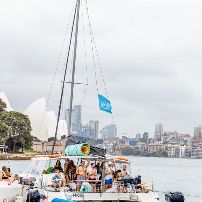 A group of people are gathered on a sailboat with a vibrant blue flag, floating on Sydney Harbour. The iconic Sydney Opera House and city skyscrapers are visible in the background under a cloudy sky. Enjoy the moment with *The Yacht Social Club* for an unforgettable experience in Luxury Yacht Rentals Sydney.