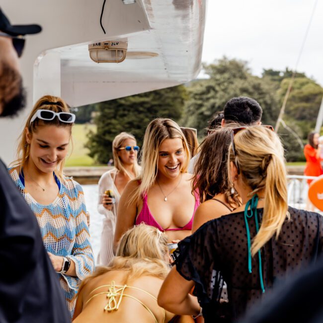 A group of people gathered on a yacht, some in swimsuits and others in casual summer clothing. Trees and water can be seen in the background. One person in a pink bikini is smiling and looking at others who are chatting. The Yacht Social Club Sydney Boat Hire logo is in the corner.