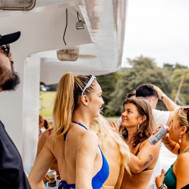 A group of people socializing on a yacht. One woman in a blue bikini and white skirt is smiling, holding a drink. Another person in sunglasses and a hat is at the left. Others in swimwear are holding drinks and chatting. The background shows greenery and the Luxury Yacht Rentals Sydney logo.