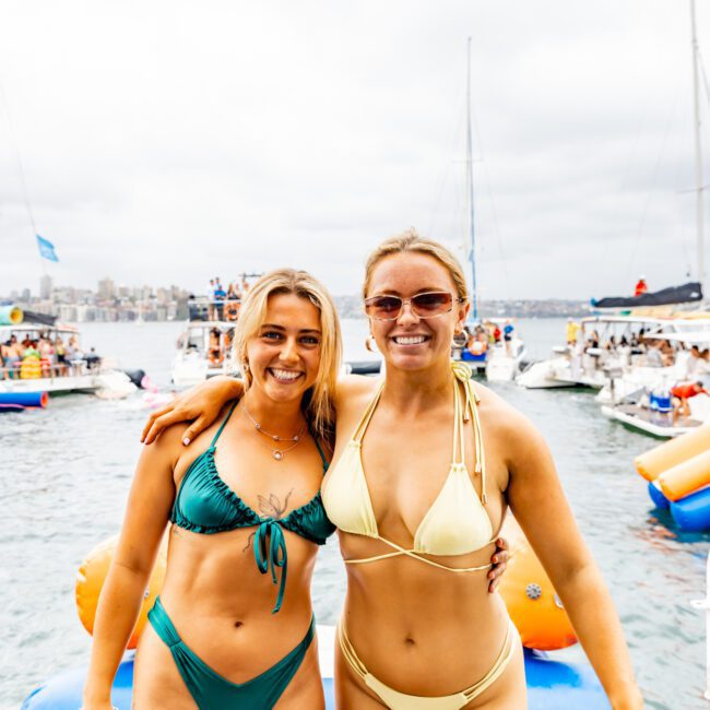 Two women in bikinis are smiling and standing side by side on a dock with arms around each other. The woman on the left is wearing a green bikini, and the woman on the right is wearing a yellow bikini. Several boats and people are visible on the water in the background, perfect for The Yacht Social Club events in Sydney.