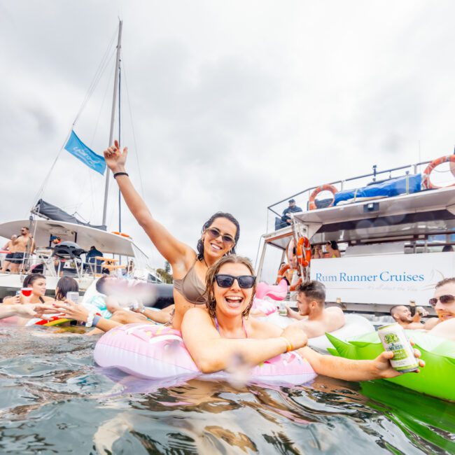 A large group of people enjoying a boat party float on an open body of water. Two women in the foreground are in inflatable pool floats—one holding a drink and smiling, the other raising her arm in excitement. A boat with "The Yacht Social Club Sydney Boat Hire" is in the background.