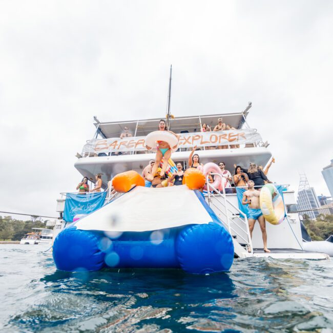 A lively group of people enjoy a day on a large party boat named "Career Explorer." Some are standing on the boat's deck while others are on an inflatable slide attached to the boat, ready to splash into the water. The background reveals a cityscape with tall buildings, embodying the essence of Luxury Yacht Rentals Sydney.