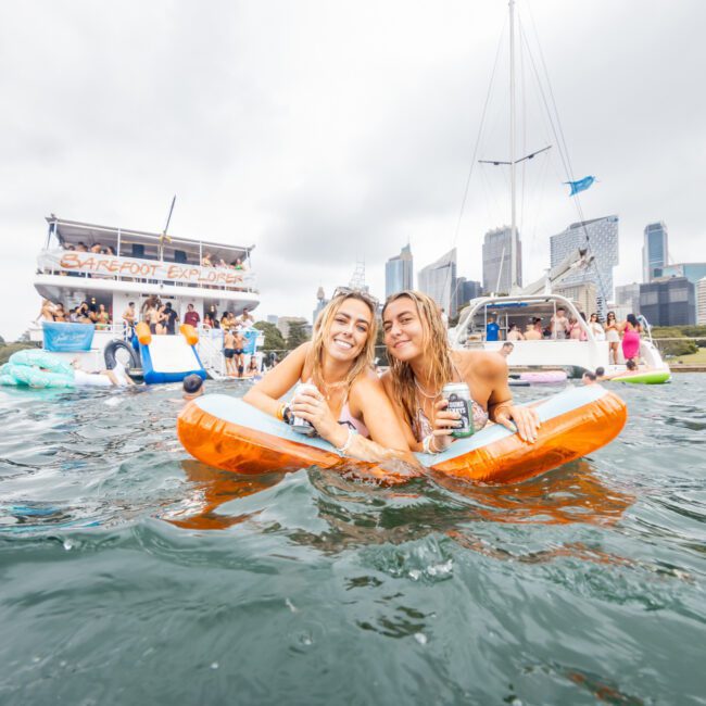 Two women smiling and holding drinks float on inflatable pool loungers in the water, with a boat labeled "Barefoot Explorer" and a sailboat nearby. City skyscrapers and trees are visible in the background, indicating a festive outdoor gathering organized by The Yacht Social Club.