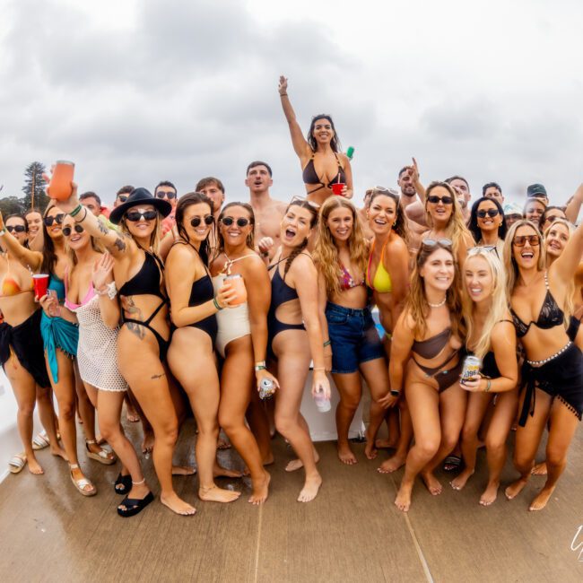 A lively group of young adults in swimwear posing on a boat, many holding drinks and smiling at the camera. The Yacht Social Club adds to the fun atmosphere as the boat is docked near a body of water with trees in the background. Some are making energetic gestures.
