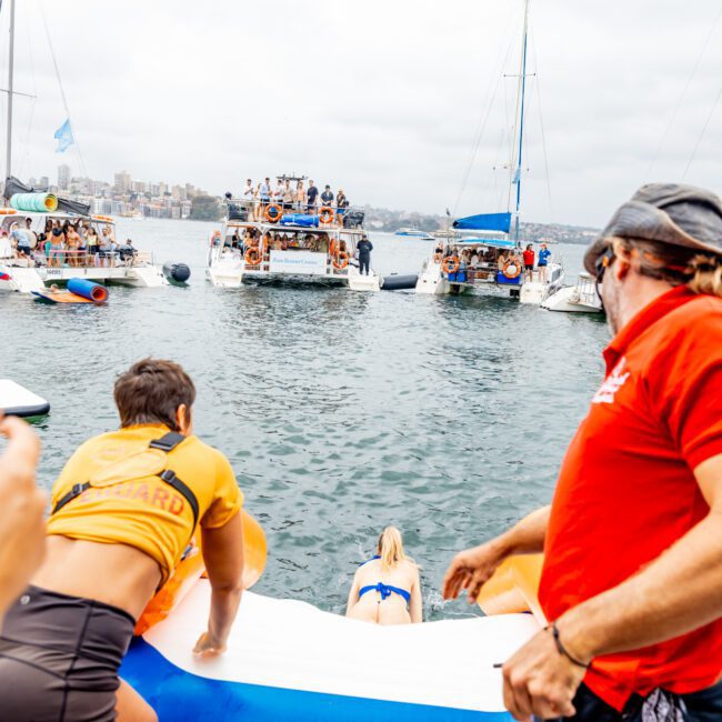 A group of people on boats and a floating platform at a lively gathering on the water. Some are watching while others enjoy inflatable structures. Two individuals in lifeguard attire oversee the activity, ensuring safety as a person slides down an inflatable ramp during The Yacht Social Club event.