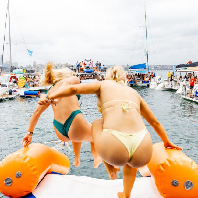 Two people in swimsuits jump into the water from an orange inflatable structure, holding hands. They are in a busy marina with several boats and many people in the background, near The Yacht Social Club Sydney Boat Hire. The sky is cloudy, and the atmosphere appears lively and festive.