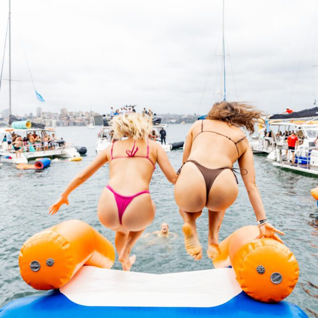 Two women wearing bikinis jump into the water from an inflatable dock platform. Boats and people are in the background on a cloudy day in Sydney Harbour, creating a lively atmosphere typical of The Yacht Social Club's events.