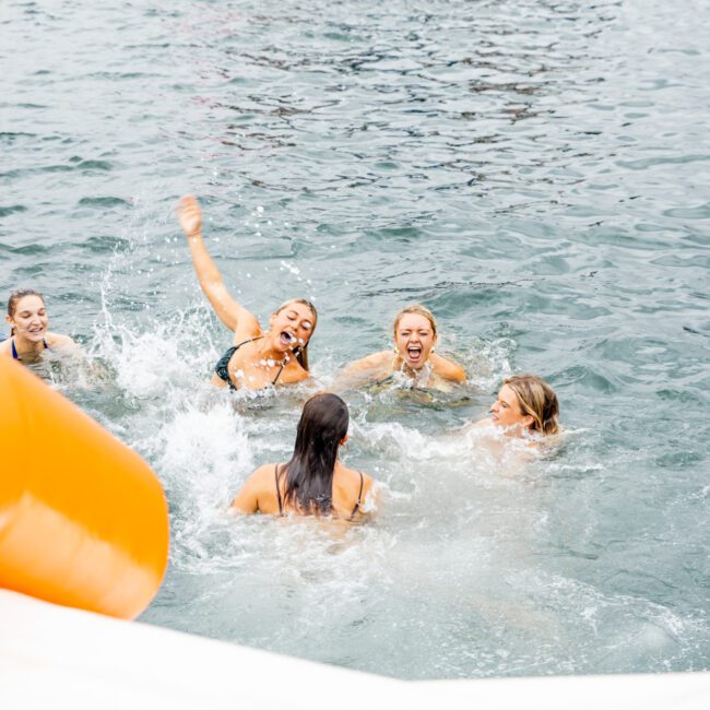 A group of people in swimsuits are in the water, joyfully splashing and laughing. In the background, there is a boat with more people enjoying inflatable pool toys. The scene suggests a fun, lively atmosphere, likely during a summer outing with The Yacht Social Club Sydney Boat Hire.