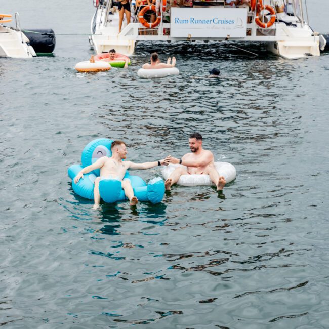 Two people float on inflatable loungers, holding hands in the water. Nearby, a group enjoys a double-deck party boat with a "Rum Runner Cruises" sign. Others float on inflatables, all with city buildings in the background. Experience this with The Yacht Social Club Event Boat Charters for an unforgettable day.