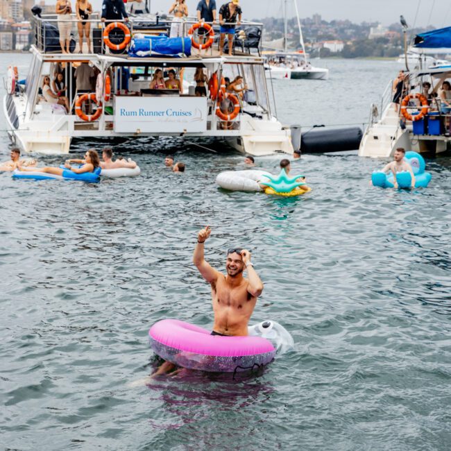 A man wearing a pink inflatable ring dances in the water near anchored yachts. People are onboard the yachts and some are in the water on floats. The left yacht has a sign "Rum Runner Cruises," hosting The Yacht Social Club event. The lively scene unfolds under a cloudy sky in the background.