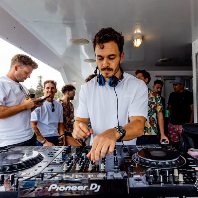 A DJ is performing on a boat from The Yacht Social Club Sydney Boat Hire, wearing headphones and adjusting controls on a Pioneer DJ setup. Several people in casual clothing are around him, some observing and others engaged in conversation. The setting appears relaxed, with the backdrop showing water and greenery.