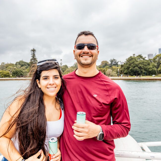 A woman with long dark hair and a man wearing sunglasses stand together on a boat, holding canned drinks. The man is wearing a red outfit, and they both smile at the camera. Trees and water are in the background, suggesting they are at The Yacht Social Club Event Boat Charters near a park.