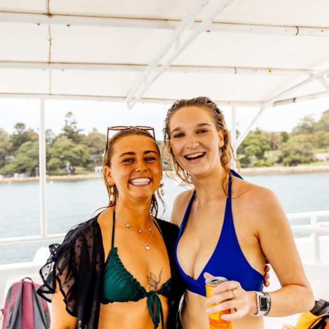 Two smiling women in swimwear pose together on a luxury yacht. One in a green bikini holds a can, while the other in a blue bikini holds a bottled drink. Both are wet, suggesting they've been swimming. Trees and water are visible in the background, embodying the perfect Sydney Harbour boat hire experience with The Yacht Social Club.