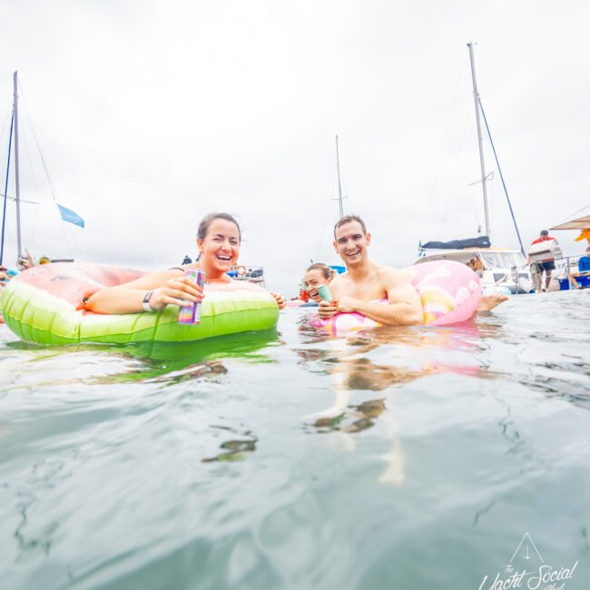 A group of people floating on colorful inflatables in a body of water, smiling and enjoying drinks with boats visible in the background. The scene appears overcast, and "The Yacht Social Club Sydney Boat Hire" logo is in the lower right corner.