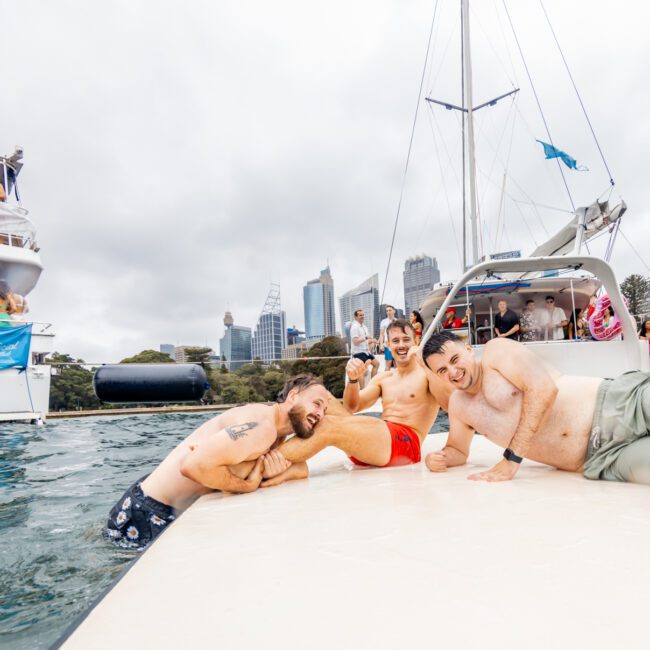 A lively scene on the water shows several men posing playfully on a floating dock, with two boats in the background filled with people enjoying a party from The Yacht Social Club. The city skyline, possibly Sydney Harbour, is visible under a cloudy sky, adding to the festive atmosphere.