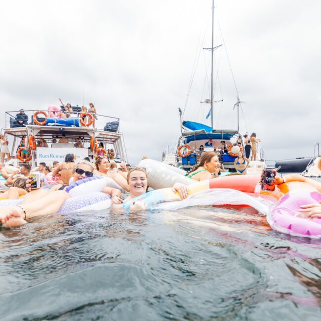 A vibrant scene on the water shows a group of cheerful people enjoying a day out on inflatable floats, including a flamingo and a unicorn. The background features several boats with more people, all part of Boat Parties Sydney The Yacht Social Club. The atmosphere is lively and festive under an overcast sky.