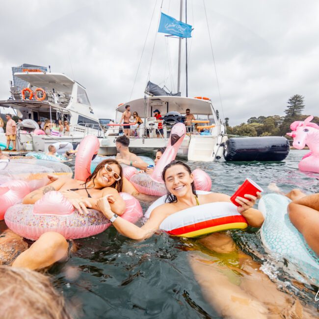 A group of people enjoying a boat party on the water, some lounging on inflatable unicorn and flamingo floats, holding drinks and smiling. In the background, there are two boats with more people and a cityscape with tall buildings. Experience unforgettable moments with Boat Parties Sydney The Yacht Social Club.
