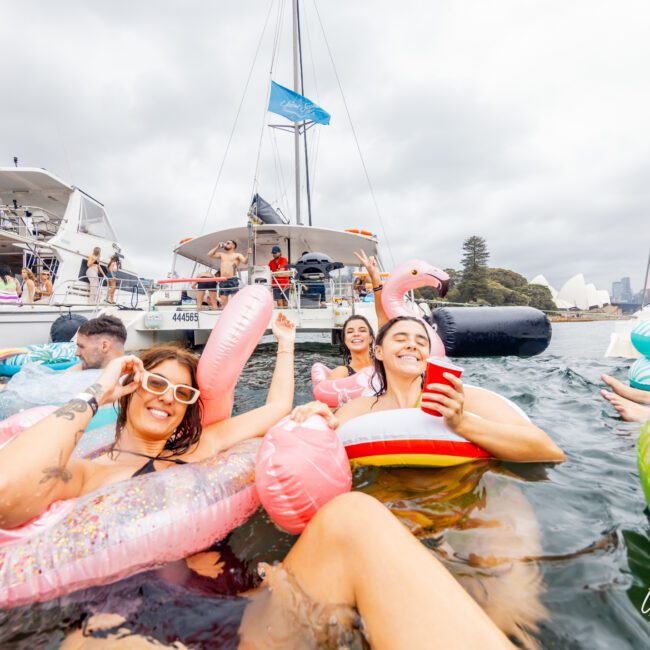 A group of people are enjoying a lively boat party in Sydney Harbour, floating on inflatable toys shaped like a flamingo, unicorn, and watermelon slice. They are smiling, holding drinks, and surrounded by various boats from The Yacht Social Club. The scene is festive and joyful.
