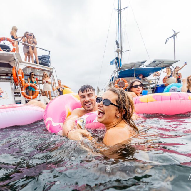People are having fun in the water, surrounded by inflatables, near some boats. Two people, a man and a woman, are smiling and holding a drink as they float on a pink inflatable. Others are swimming and relaxing nearby, enjoying the lively atmosphere of The Yacht Social Club.