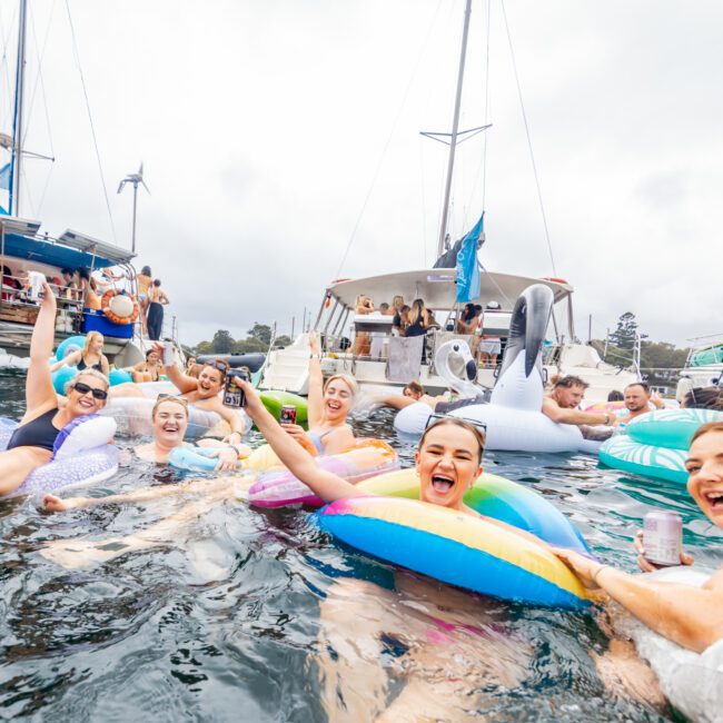 A lively group of people are seen swimming and relaxing on bright inflatable tubes in the water, with several yachts from The Yacht Social Club Sydney Boat Hire anchored nearby. They appear to be having a joyful time, some holding drinks and laughing. The sky is overcast, and the mood is festive and fun.
