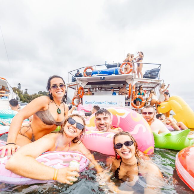 A group of people enjoying a sunny day in the water with inflatable floaties. They are smiling and posing for a photo in front of a boat labeled "Rum Runner Cruises," which embodies the charm often found with Luxury Yacht Rentals Sydney. The boat has a few more people relaxing on it as the sky is partly cloudy.