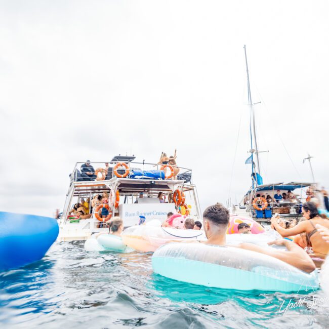 People floating on inflatable rafts in the water near two anchored boats, enjoying a fun day under a cloudy sky. The larger boat has "Rum Runner Cruise" written on it and people are gathered on the deck. The scene appears lively and festive, embodying the spirit of Boat Parties Sydney by The Yacht Social Club.
