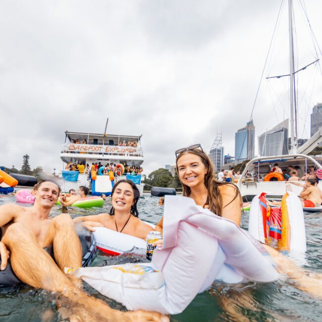A group of people floating on inflatable rafts in the water, surrounded by boats and other rafts, enjoying a fun time. A city skyline with tall buildings is visible in the background. The Yacht Social Club events perfectly capture this lively day on the water in Sydney.